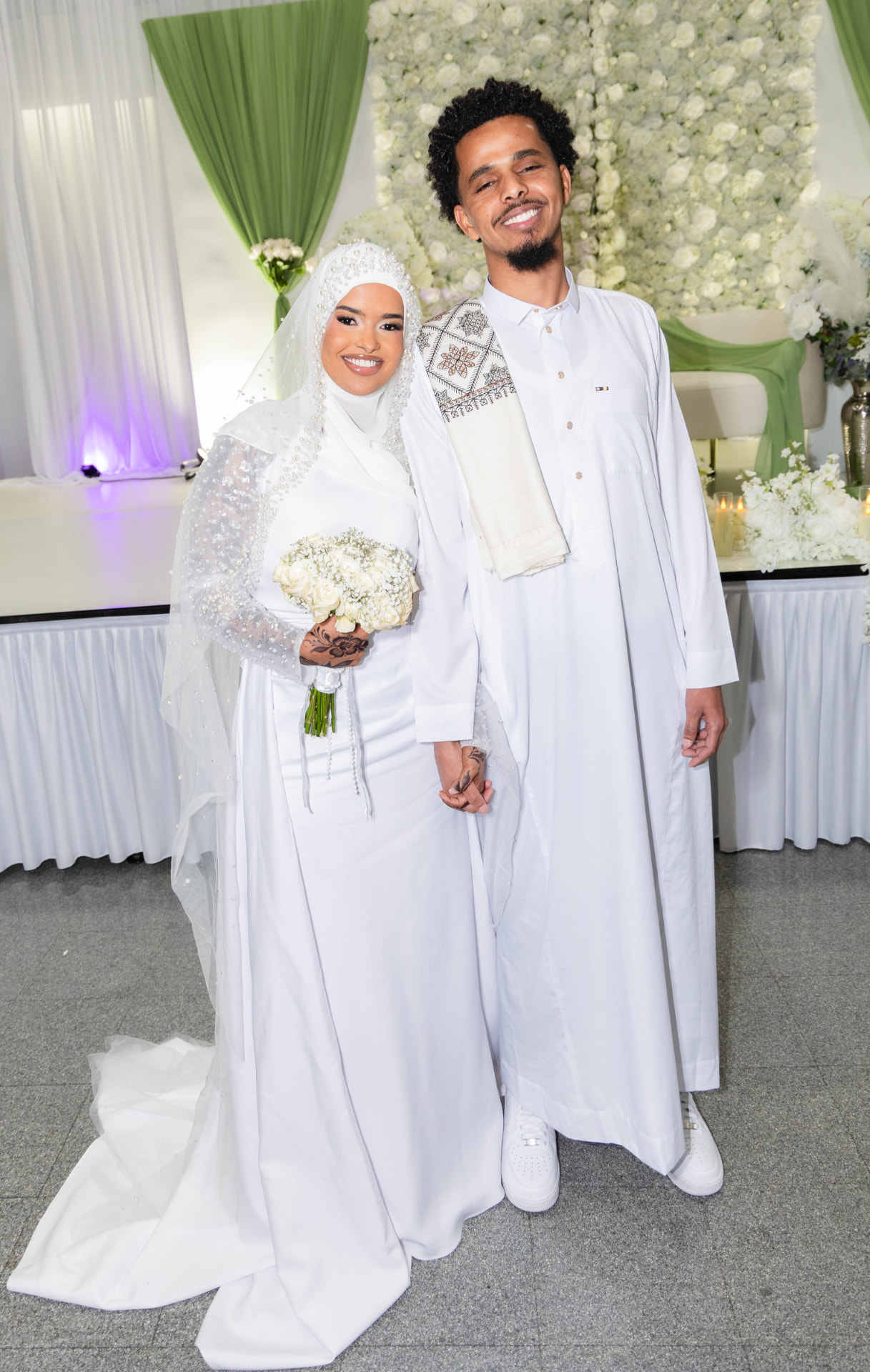 Bride and groom in white before a white rose flower wall at their nikah