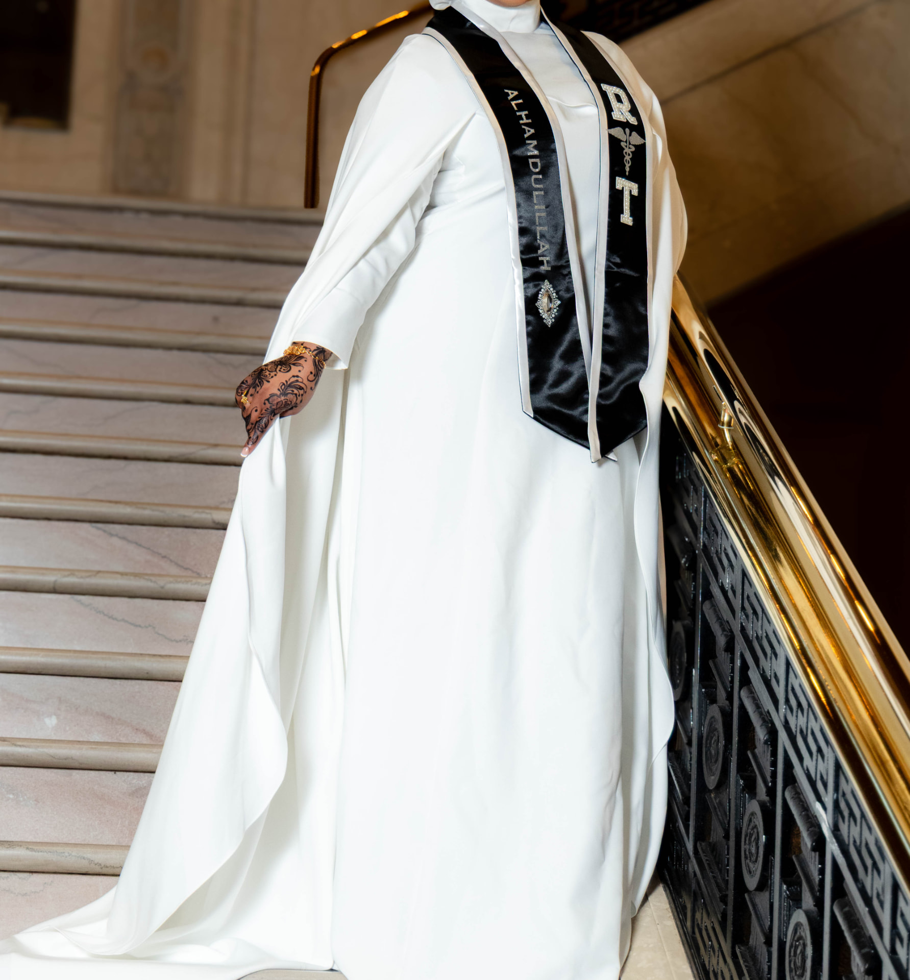 Graduate in a white gown and black honors stole with henna detailing on the capitol staircase
