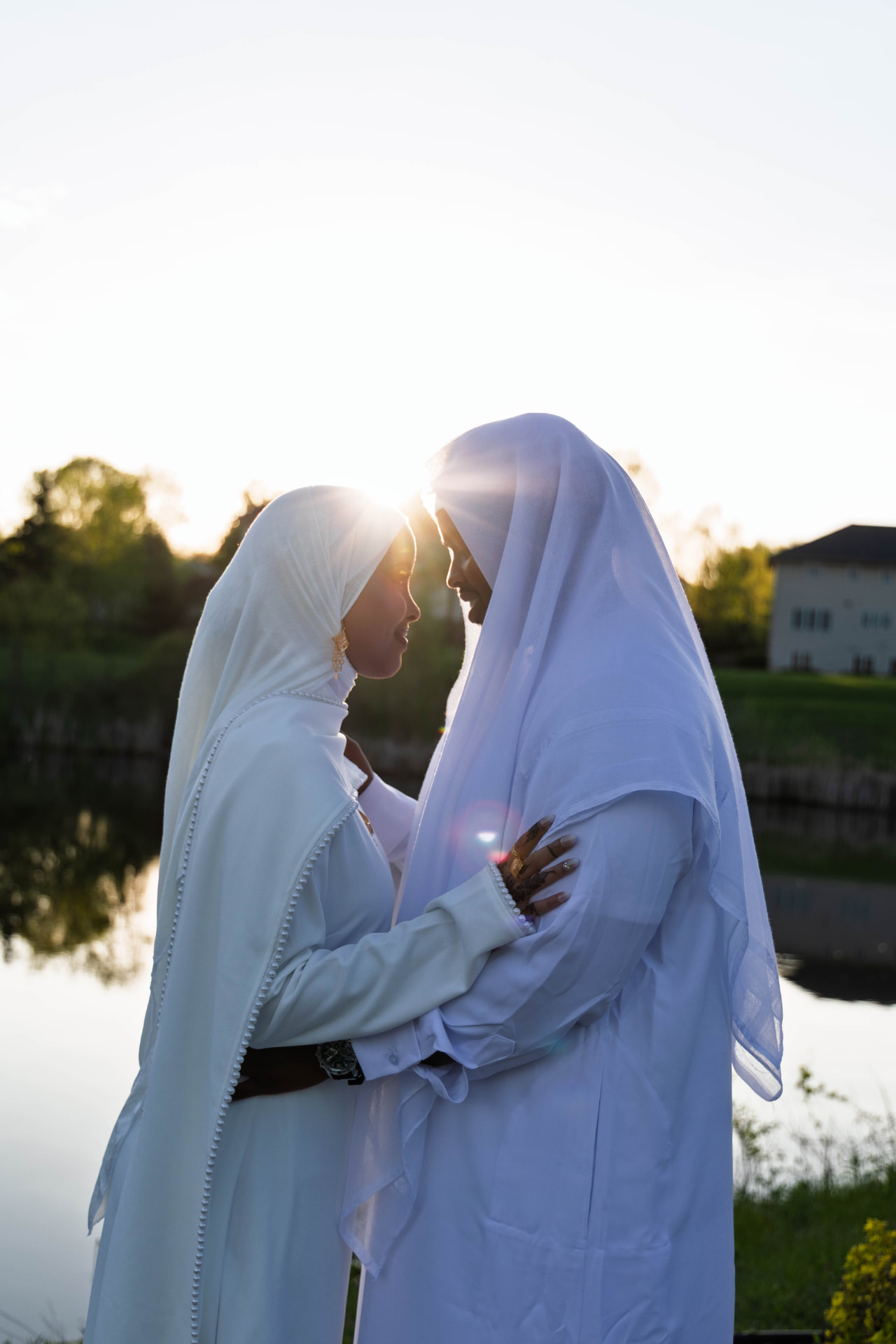 Couple in white nikkah attire forehead to forehead at golden hour by a lake