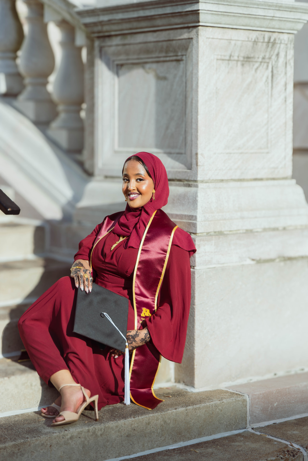 Graduate in maroon regalia seated on capitol steps holding her cap