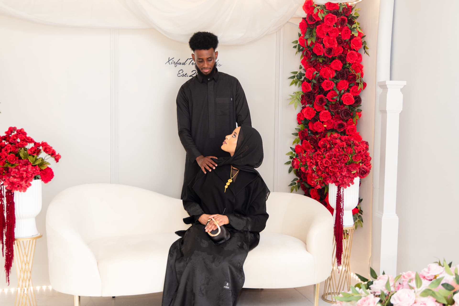 Couple in black attire seated on a cream sofa beside a red rose wall at Xirfad Studio
