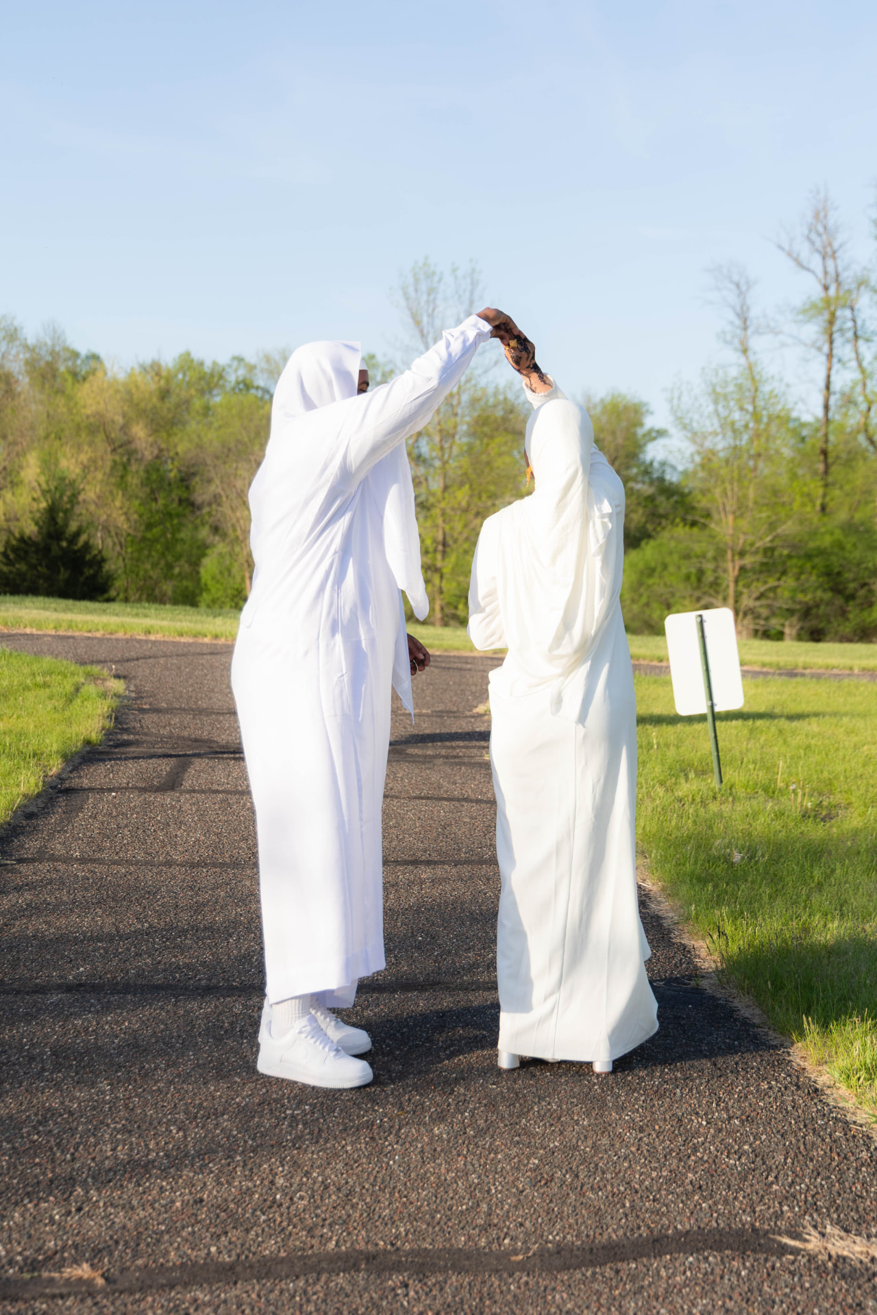 Groom twirling his bride, both in white, on a sunlit country path
