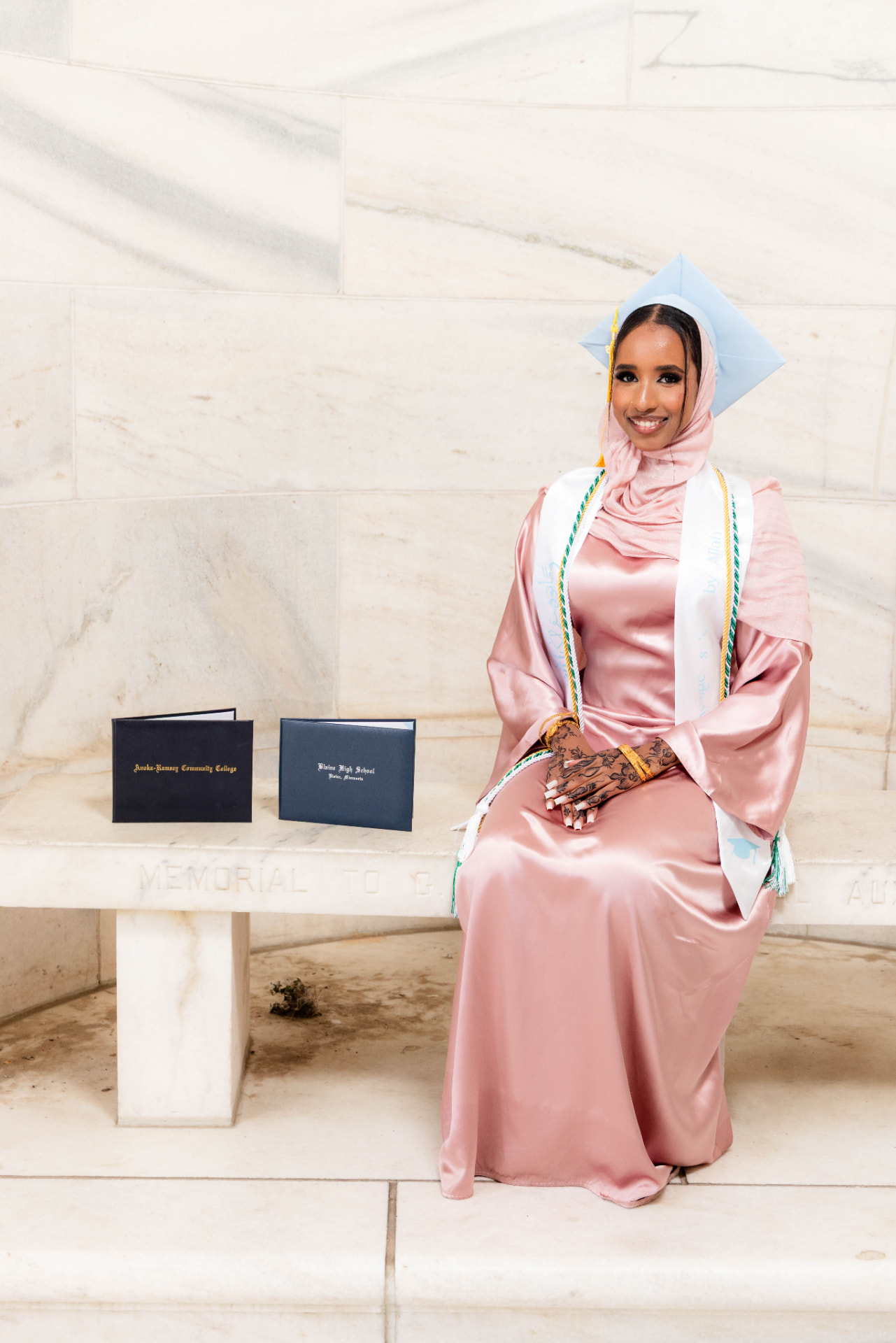 Graduate in blush satin seated with two diplomas on a marble bench