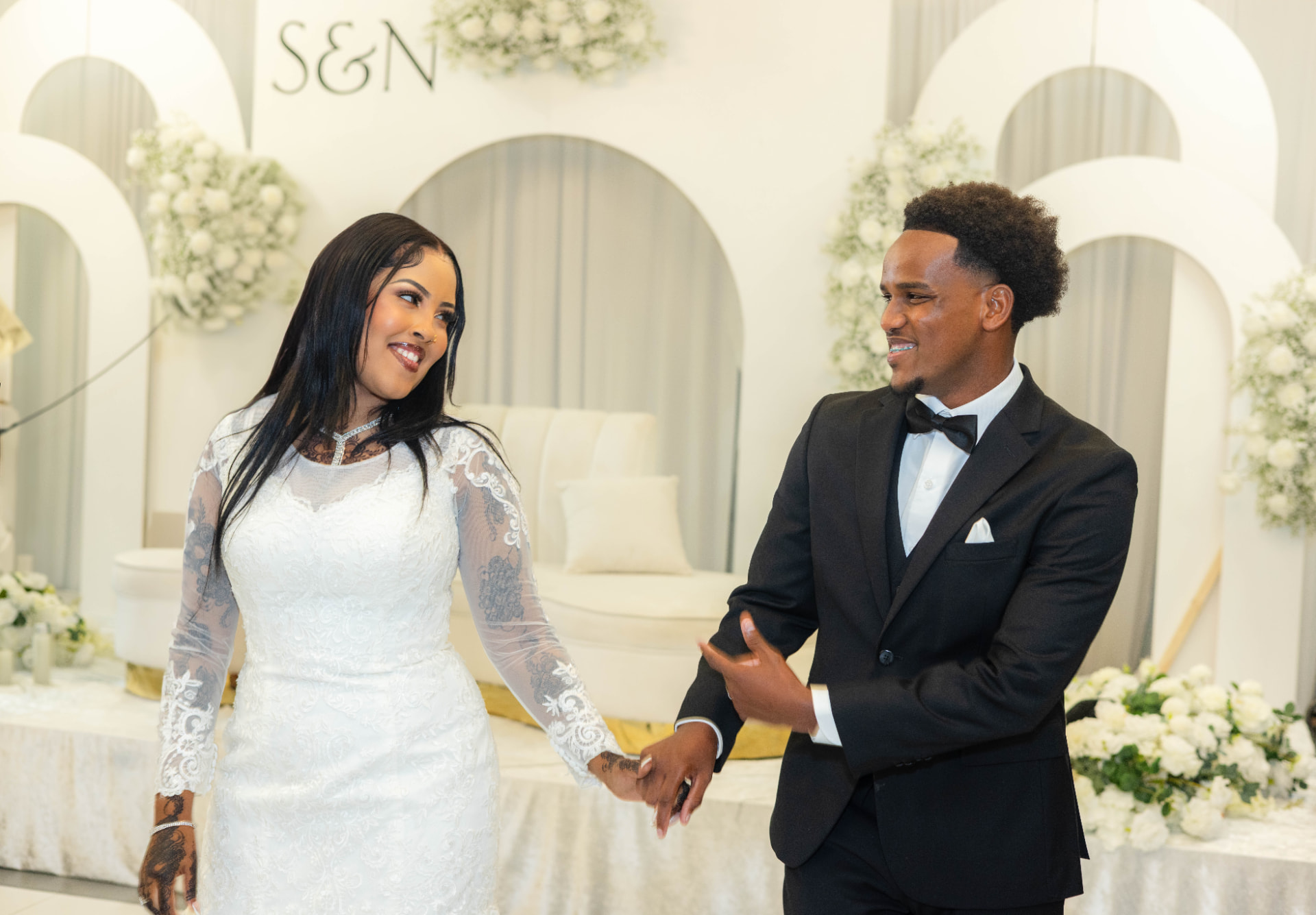 Bride in white lace and groom in a black tuxedo smiling before an ivory arch reception backdrop