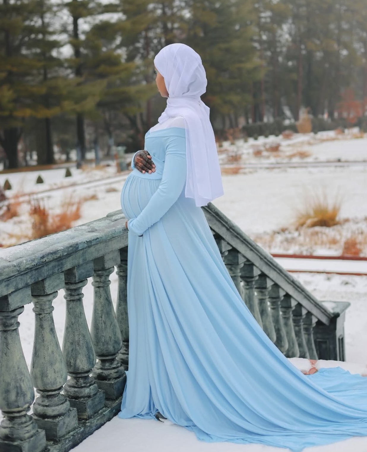 Expecting mother in a powder-blue maternity gown cradling her bump beside a stone balustrade in the snow