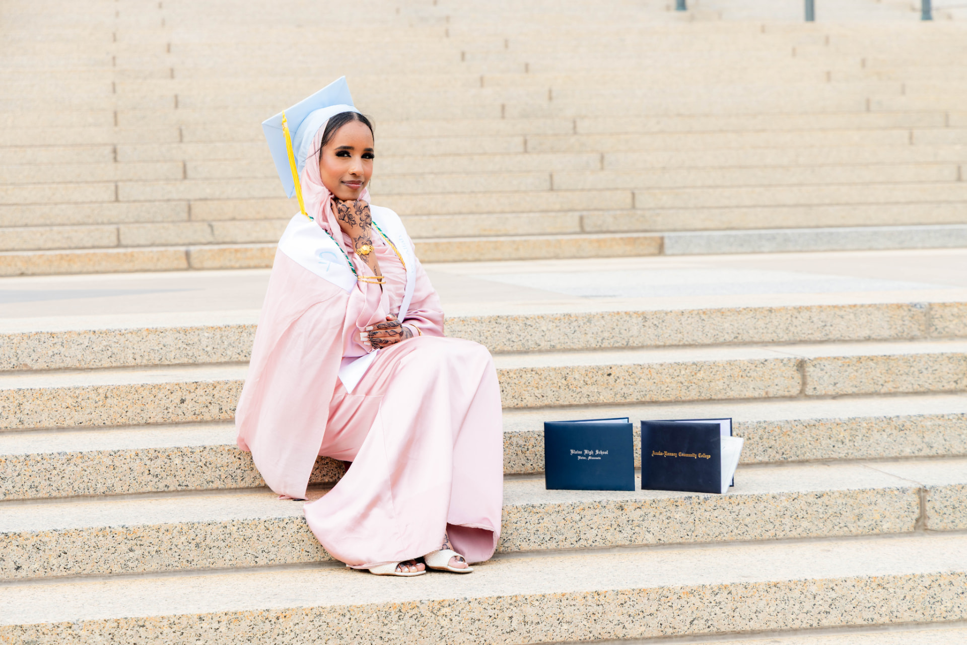 Graduate in blush and light blue regalia seated beside two diplomas on stone steps