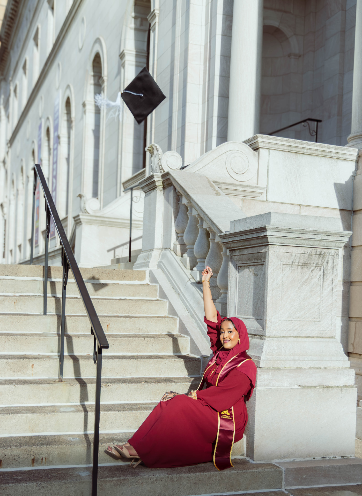 Graduate in maroon University of Minnesota regalia tossing her cap on the capitol steps
